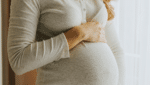 Why Summer is the Perfect Time to Explore Surrogacy with Conceptual Options CO Blog Post Image 061026 - Conceptual Options Surrogacy Agency and Egg Donation Agency Pregnant woman cradling her baby bump with one hand over her abdomen, standing near a window in a light-gray knit top with lace trim at the neckline.