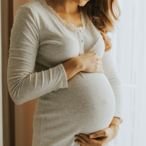 Why Summer is the Perfect Time to Explore Surrogacy with Conceptual Options CO Blog Post Image 061026 - Conceptual Options Surrogacy Agency and Egg Donation Agency - Conceptual Options Surrogacy Agency and Egg Donation Agency Pregnant woman cradling her baby bump with one hand over her abdomen, standing near a window in a light-gray knit top with lace trim at the neckline.